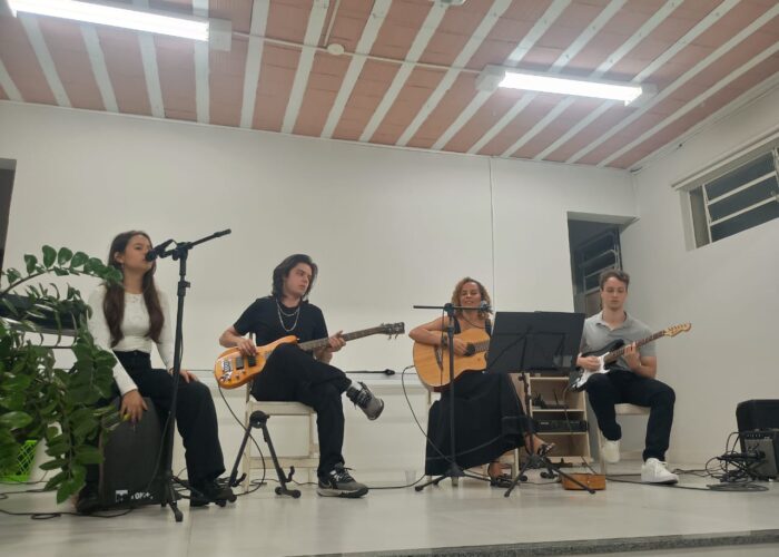 A esquerda mulher pele branca cabelo preto, camisa branca, calça preta, sentada tocando cájon, homem branco, camisa e calça preta, sentado tocando guitarra, mulher branca cabelo loiro, vestido preto, sentada tocando violão, homem branco, cabelo preto, camisa cinza, calça preta, sentado tocando guitarra.