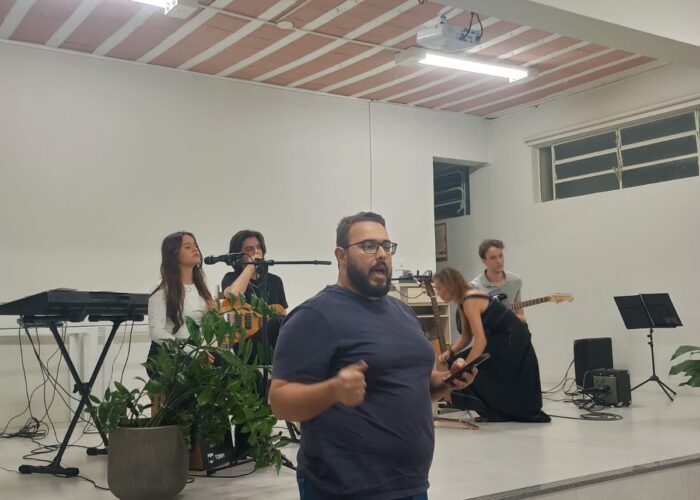 A frente homem branco, cabelo e barba preta, óculos, camisa escura, calça jeans, em pé, ao fundo, a esquerda mulher pele branca cabelo preto, camisa branca, calça preta, sentada tocando cájon, homem branco, camisa e calça preta, sentado tocando guitarra, mulher branca cabelo loiro, vestido preto, sentada tocando violão, homem branco, cabelo preto, camisa cinza, calça preta, sentado tocando guitarra.