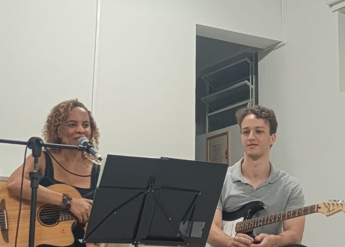 A esquerda mulher branca cabelo loiro, vestido preto, sentada tocando violão, homem branco, cabelo preto, camisa cinza, calça preta, sentado tocando guitarra.