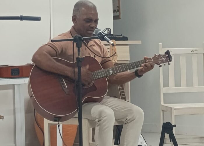 Homem pele negra camisa marrom, sentado tocando violão