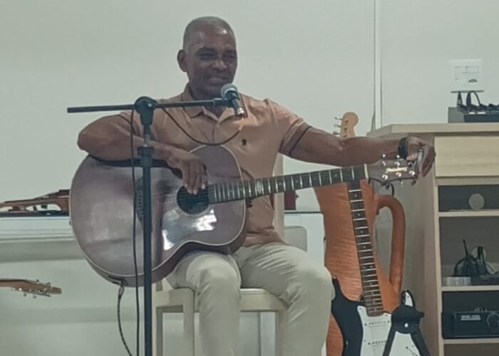 Homem pele negra camisa marrom, sentado tocando violão