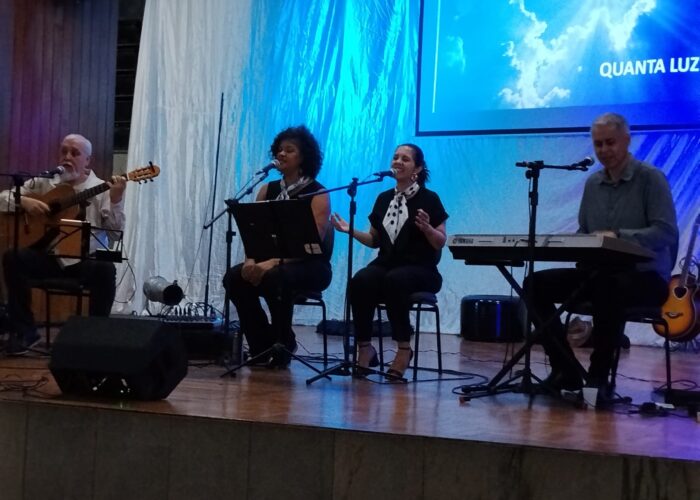 A esquerda homem pele branca, cabelo e barba branca, camisa branca, sentado tocando violão, mulher negra cabelo preto camisa preta, sentada cantando, mulher branca, cabelo preto, camisa preta, sentada cantando, homem branco cabelo grisalho, camisa cinza, sentado tocando teclado.