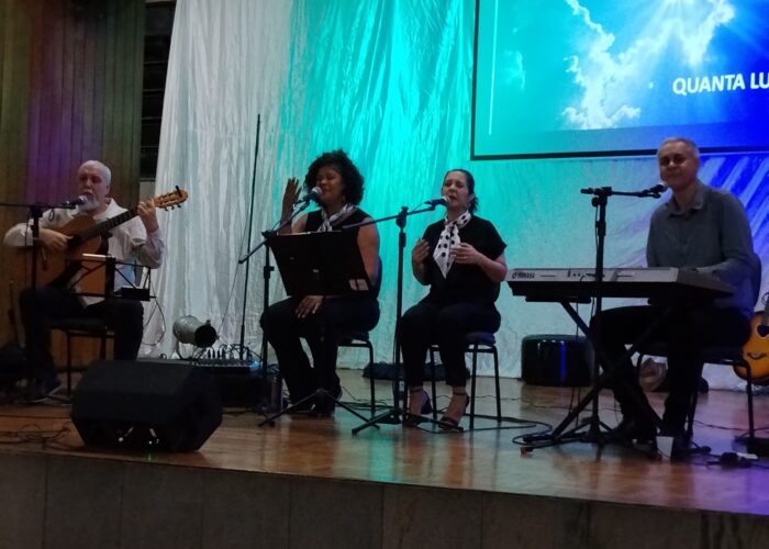A esquerda homem pele branca, cabelo e barba branca, camisa branca, sentado tocando violão, mulher negra cabelo preto camisa preta, sentada cantando, mulher branca, cabelo preto, camisa preta, sentada cantando, homem branco cabelo grisalho, camisa cinza, sentado tocando teclado.