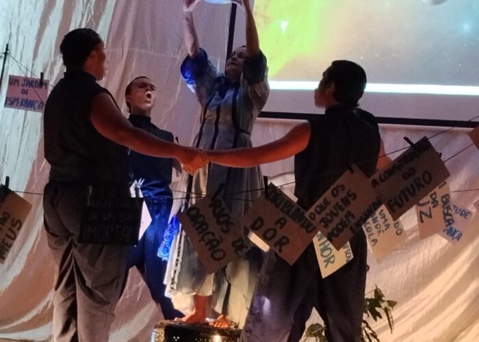 No centro mulher branca, vestido azul, segurando ao alto um globo terrestre, ao redor da mulher quatro pessoas com roupas pretas, de mãos dadas formando um circulo.
