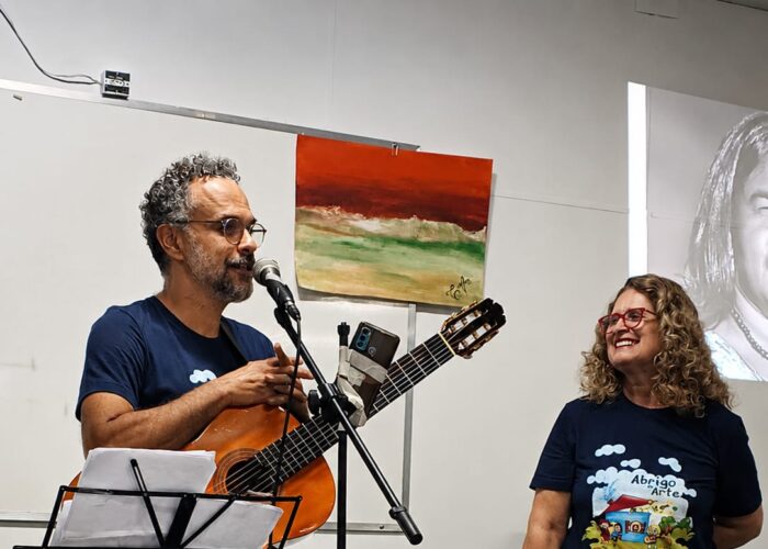 A esquerda homem branco, cabelo grisalho, óculos, camisa azul, calça verde escuro, segurando violão, mulher branca, cabelo loiro, óculos, camisa preta, calça preta.