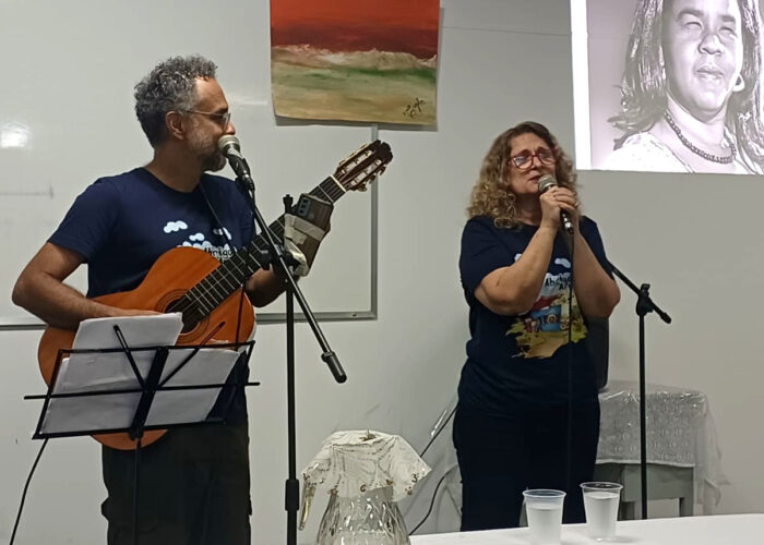 A esquerda homem branco, cabelo grisalho, óculos, camisa azul, calça verde escuro, segurando violão, mulher branca, cabelo loiro, óculos, camisa preta, calça preta, cantando no microfone.
