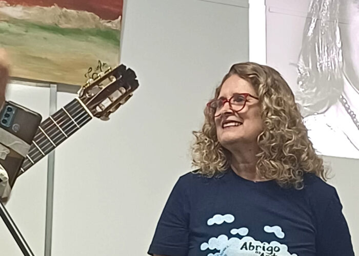 Mulher branca, cabelo loiro, óculos, camisa preta, calça preta.