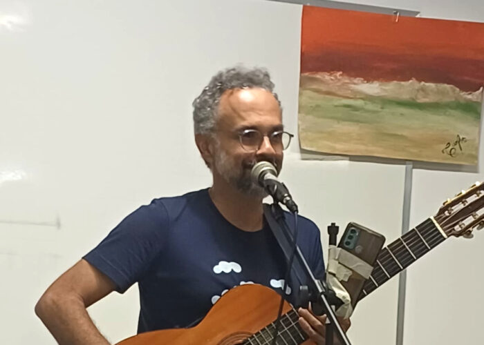 Homem branco, cabelo grisalho, óculos, camisa azul, calça verde escuro, segurando violão.