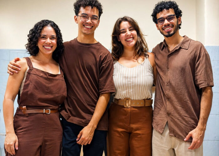 A esquerda mulher pele branca, cabelo preto, camisa e calça marrom, homem pele branca, cabelo preto, óculos, camisa marrom, calça preta, mulher pele branca, cabelo loiro, camisa branca, calça marrom, homem pele branca, cabelo preto, óculos, camisa marrom, calça bege.