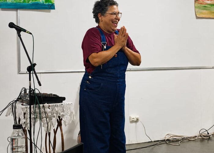 Mulher branca sorrindo, cabelo preto, óculos, camisa vinho, macacão azul.