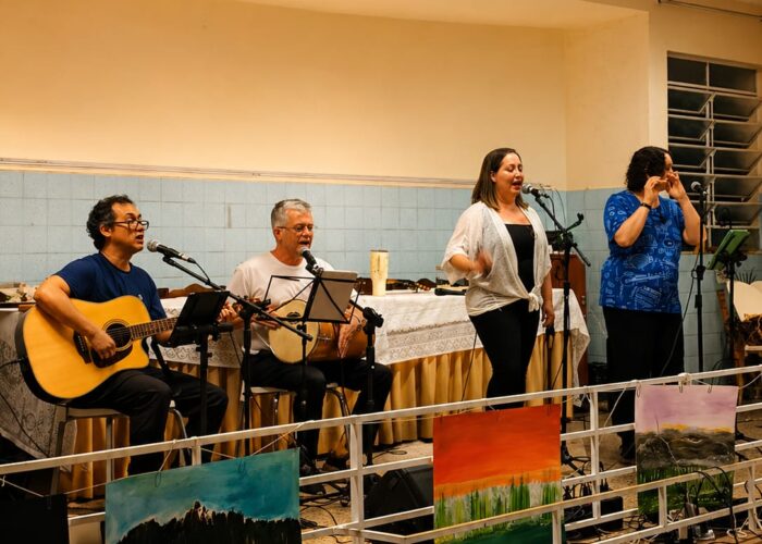 A esquerda homem pele branca, cabelo preto, óculos, camisa azum, calça preta, sentado tocando violão, homem pele branca, cabelo branco, camisa branca, calça preta, sentado tocando tambor, mulher pele branca, cabelo loiro, camisa branca, calça preta, em pé cantando, mulher pele branca, cabelo preto, camisa azul, calça preta, em pé tocando flauta.