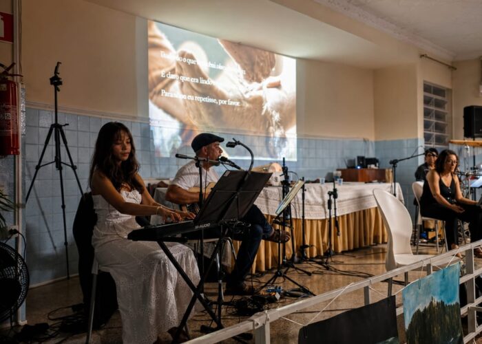 A esquerda mulher pele branca, cabelo preto, vestido branco, sentada tocando teclado, homem pele branca, boné, camisa branca, calça jeans, sentado tocando violão, mulher pele branca, cabelo preto, camisa e calça preto, sentada cantando, homem pele branca, gorro preto, camisa e calça jeans, sentado.