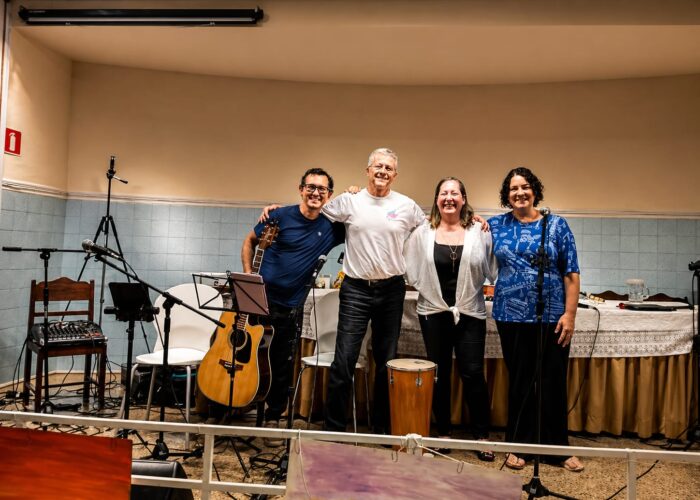 A esquerda homem pele branca, cabelo preto, óculos, camisa azum, calça preta, segurando violão, homem pele branca, cabelo branco, camisa branca, calça preta, mulher pele branca, cabelo loiro, camisa branca, calça preta, mulher pele branca, cabelo preto, camisa azul, calça preta.