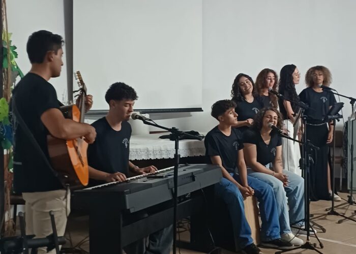 A esquerda rapaz pele parda, cabelo preto, camisa preta, calça bege, em pé tocando violão, rapaz pele branca, cabelo preto, camisa preta, sentado tocando teclado, rapaz pele branca, cabelo preto, camisa preta, calça jeans, sentado tocando Cajon, moça pele branca, cabelo loiro, camisa preta, calça jeans, sentada e cantando, moça pele brança, cabelo preto, camisa preta, cantando, moça pele brança, cabelo loiro, camisa preta, cantando, moça pele brança, cabelo preto, camisa preta, calça branca, em pé cantando, moça pele parda, cabelo loiro, camisa e calça preta, em pé cantando.