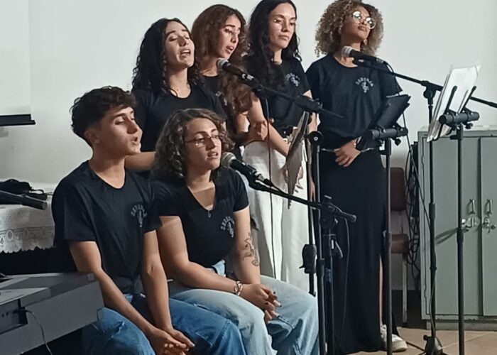 A esquerda rapaz pele branca, cabelo preto, camisa preta, calça jeans, sentado tocando Cajon, moça pele branca, cabelo loiro, camisa preta, calça jeans, sentada e cantando, moça pele brança, cabelo preto, camisa preta, cantando, moça pele brança, cabelo loiro, camisa preta, cantando, moça pele brança, cabelo preto, camisa preta, calça branca, em pé cantando, moça pele parda, cabelo loiro, camisa e calça preta, em pé cantando.