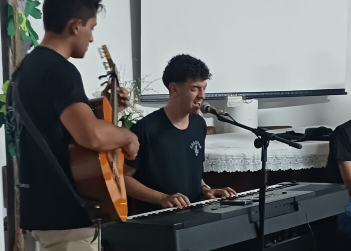 Rapaz pele parda, cabelo preto, camisa preta, calça bege, em pé tocando violão, rapaz pele branca, cabelo preto, camisa preta, sentado tocando teclado.
