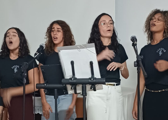 A esquerda moça pele brança, cabelo preto, camisa preta, calça marrom, em pé cantando, moça pele brança, cabelo loiro, camisa preta, calça jeans, em pé cantando, moça pele brança, cabelo preto, camisa preta, calça branca, em pé cantando, moça pele parda, cabelo loiro, camisa e calça preta, em pé cantando.