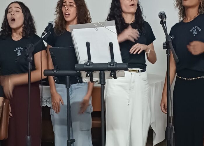 A esquerda moça pele brança, cabelo preto, camisa preta, calça marrom, em pé cantando, moça pele brança, cabelo loiro, camisa preta, calça jeans, em pé cantando, moça pele brança, cabelo preto, camisa preta, calça branca, em pé cantando, moça pele parda, cabelo loiro, camisa e calça preta, em pé cantando.