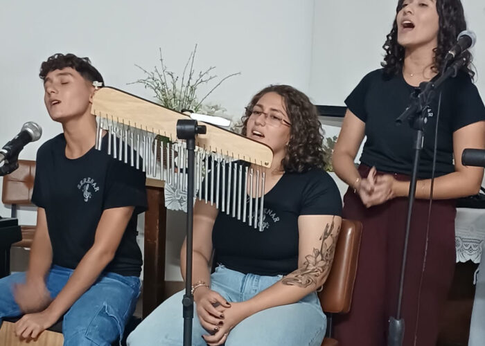 A esquerda rapaz pele branca, cabelo preto, camisa preta, calça jeans, sentado tocando Cajon, moça pele branca, cabelo loiro, camisa preta, calça jeans, sentada e cantando, moça pele brança, cabelo preto, camisa preta, calça marrom, em pé cantando.
