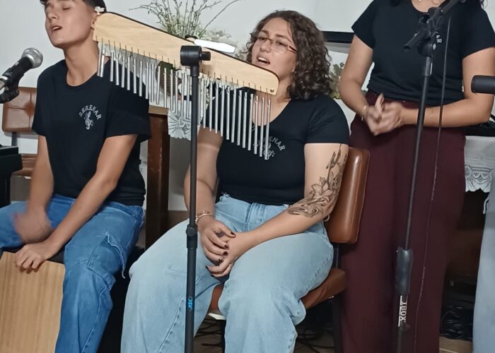 A esquerda rapaz pele branca, cabelo preto, camisa preta, calça jeans, sentado tocando Cajon, moça pele branca, cabelo loiro, camisa preta, calça jeans, sentada e cantando, moça pele brança, cabelo preto, camisa preta, calça marrom, em pé cantando.