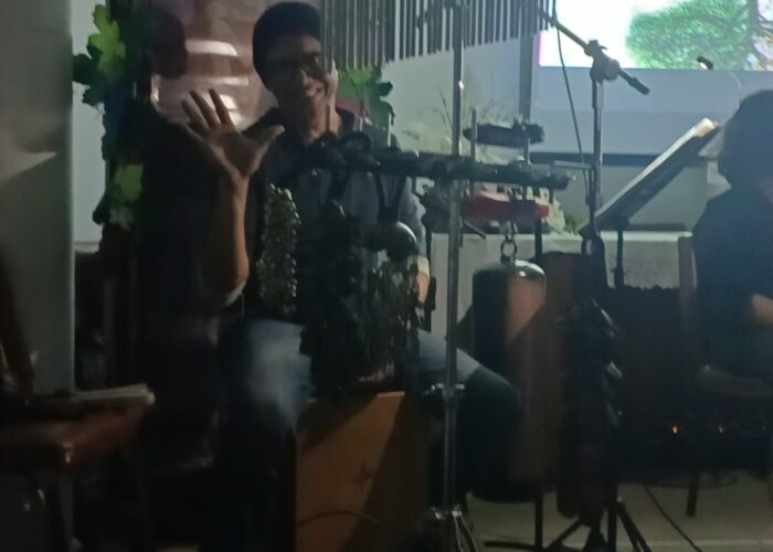 Homem pele branca, gorro preto, óculos, camisa e calça preta, sentado tocando Cajon.
