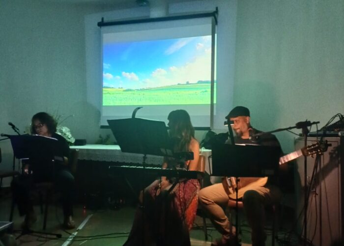 A esquerda mulher pele branca, cabelo preto, óculos, camisa e calça preta, sentada cantando, mulher pele branca, cabelo preto, vestido colorido, sentada tocando teclado, homem pele branca, boné preto, camisa preta, calça bege, sentado tocando violão.