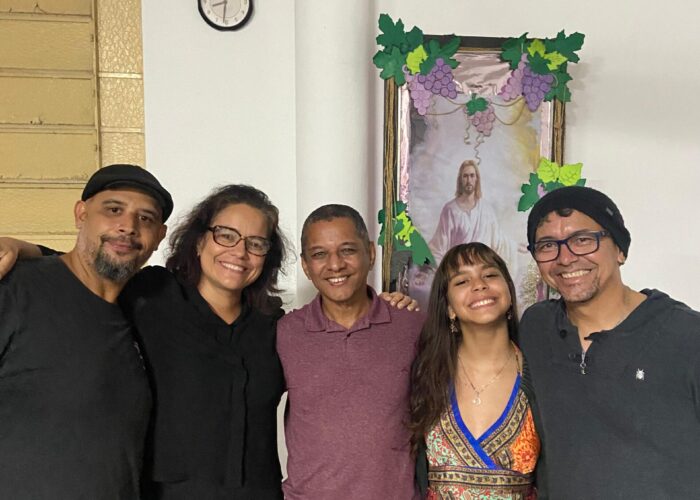 A esquerda homem pele branca, boné preto, camisa preta, calça bege, em pé, mulher pele branca, cabelo preto, óculos, camisa e calça preta, em pé, homem pele parda, cabelo preto, camisa roxa, calça bege, em pé, mulher pele branca, cabelo preto, vestido colorido, em pé, homem pele branca, gorro preto, óculos, camisa e calça preta, em pé.