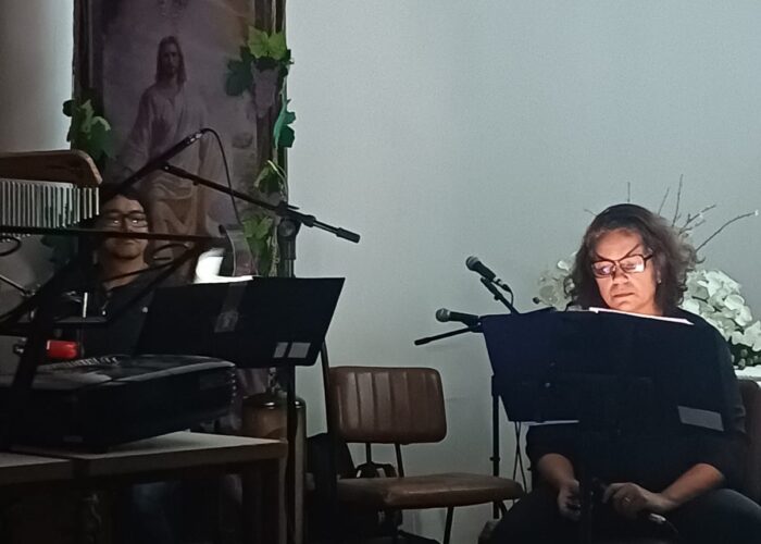 A esquerda homem pele parda, gorro preto, óculos, camisa e calça preta, sentado tocando Cajon, mulher pele branca, cabelo preto, óculos, camisa e calça preta, sentada cantando.