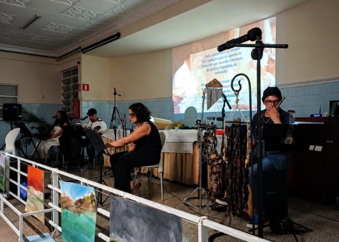 A esquerda mulher pele branca, cabelo preto, vestido branco, sentada tocando teclado, homem pele branca, boné, camisa branca, calça jeans, sentado tocando violão, mulher pele branca, cabelo preto, camisa e calça preto, sentada cantando, homem pele branca, gorro preto, camisa e calça jeans, sentado.