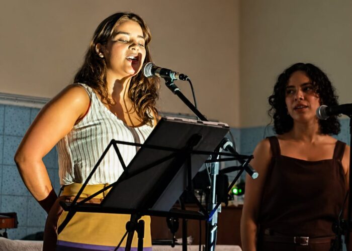 mulher pele branca, cabelo loiro, camisa branca, calça marrom, em pé cantando, mulher pele branca, cabelo preto, camisa e calça marrom, em pé cantando.