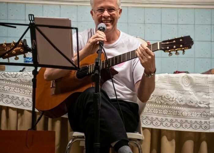 homem pele branca, cabelo branco, camisa branca, calça preta, sentado tocando violão.