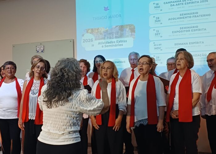 A frente mulher pele branca, cabelo grisalho, blusa branca, calca preta, atrás coral de mulheres e homens vestindo blusa branca, cachecol vermelho, calça preta.