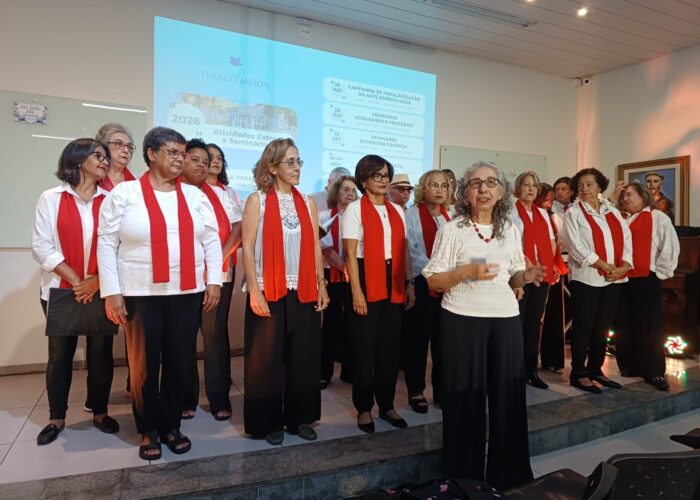 A frente mulher pele branca, cabelo grisalho, blusa branca, calca preta, atrás coral de mulheres e homens vestindo blusa branca, cachecol vermelho, calça preta.