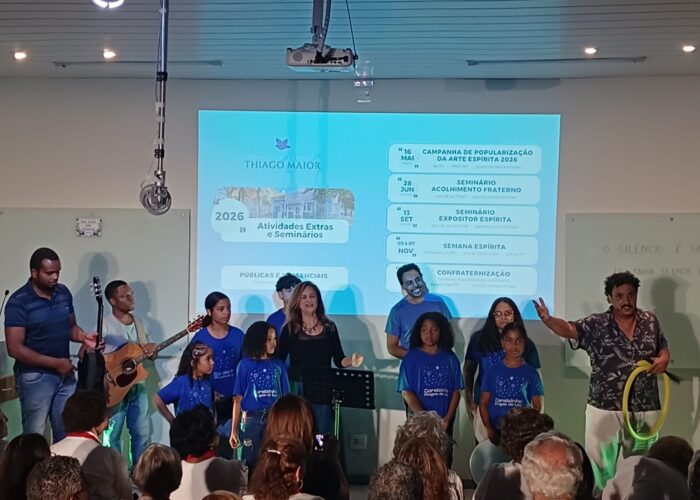 A esquerda dois homens pele preta, cabelo preto, tocando violão, crianças com camisa azul e mulher pele branca, cabelo loiro, camisa preta ao centro, homem pele parda, cabelo preto, camisa marrom, calça branca a direita.