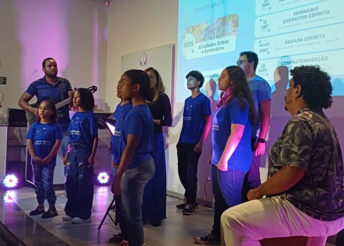 Crianças com camisa azul, calça azul cantando e homem pele parda, cabelo preto, camisa marrom, calça branca, sentado tocando cajon.