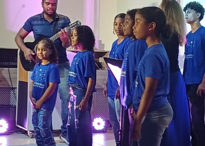 A esquerda homem pele preta, camisa azul, cabelo preto, tocando violão, crianças com camisa azul, calça azul, cantando.