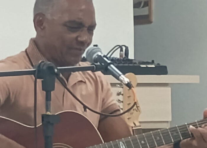 Homem pele negra camisa marrom, sentado tocando violão