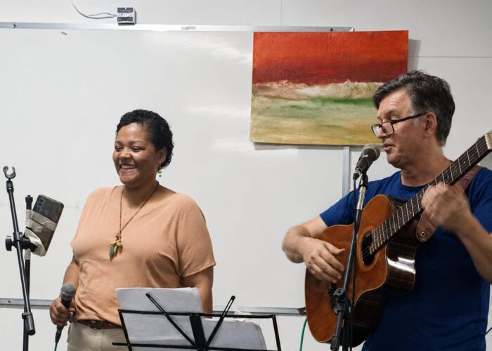 A esquerda mulher negra, cabelo preto, camisa marrom, calça cinza claro, segurando microfone, homem branco, cabelo preto, óculos, camisa azul, calçapreta, tocando violão.