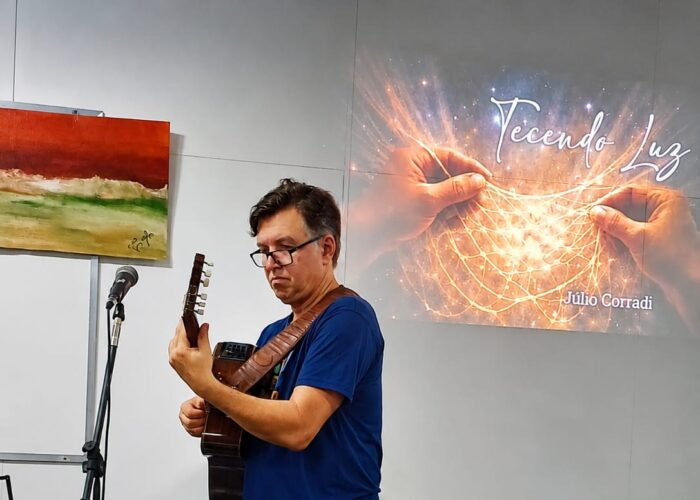 A esquerda homem branco, cabelo preto, óculos, camisa azul, calça preta, sapato marrom, tocando violão.