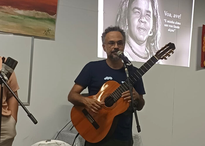 Homem branco, cabelo grisalho, óculos, camisa azul, calça verde, tocando violão.
