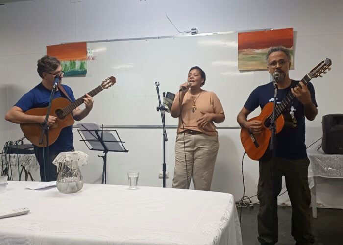 A esquerda homem branco, cabelo preto, óculos, camisa azul, calça preta, tocando violão, mulher negra, cabelo preto, camisa marrom, calça cinza claro, cantando com microfone, homem branco, cabelo grisalho, óculos, camisa azul, calça verde, tocando violão.